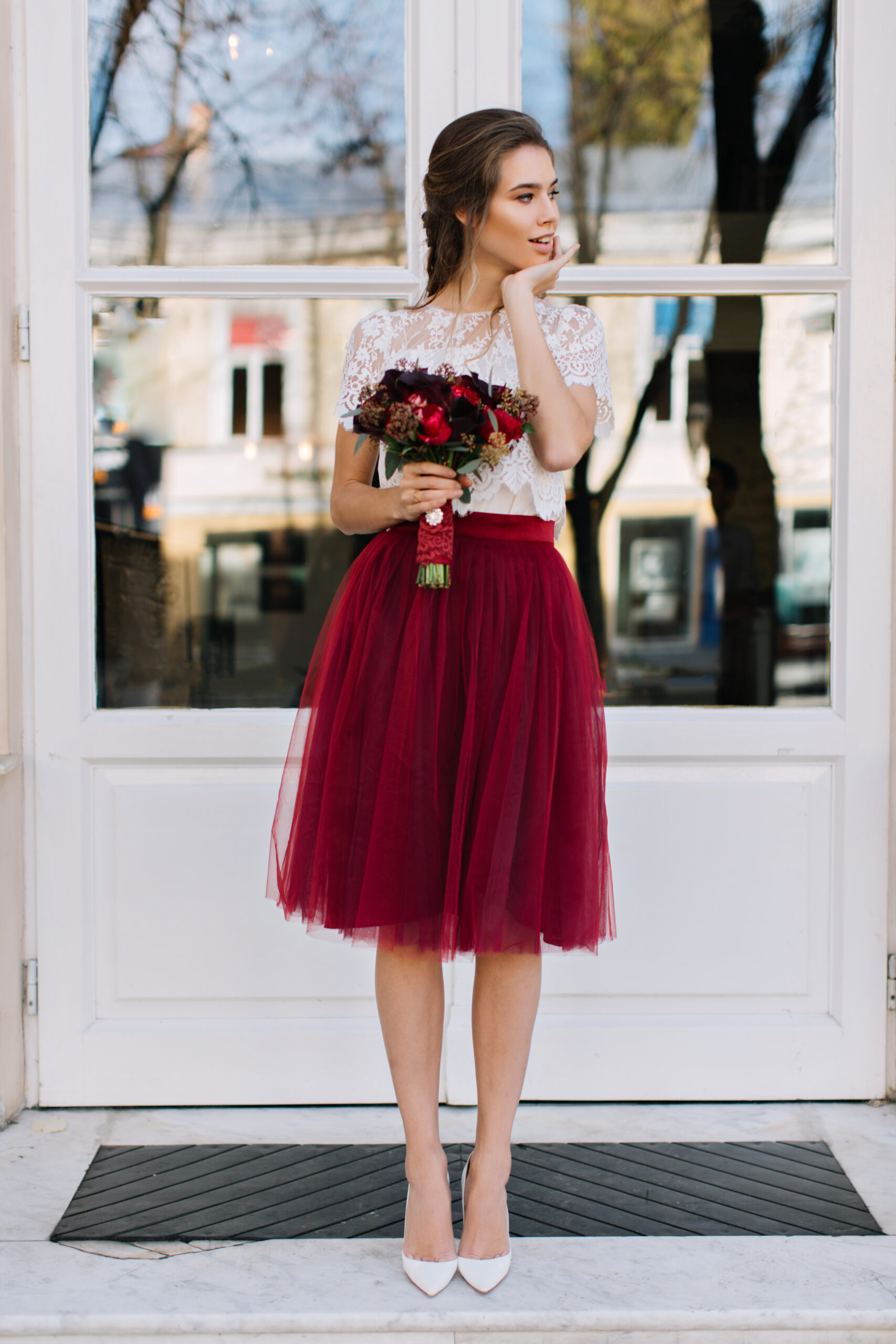 Beautiful girl in marsala tulle skirt on heels walking on street. She holds boquet of flowers and smiling to side